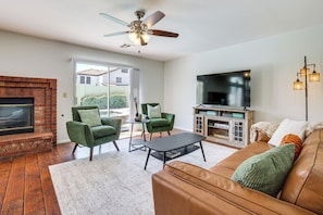 A living room with a fireplace, green armchairs, a brown leather sofa, a flat-screen TV, and a ceiling fan.