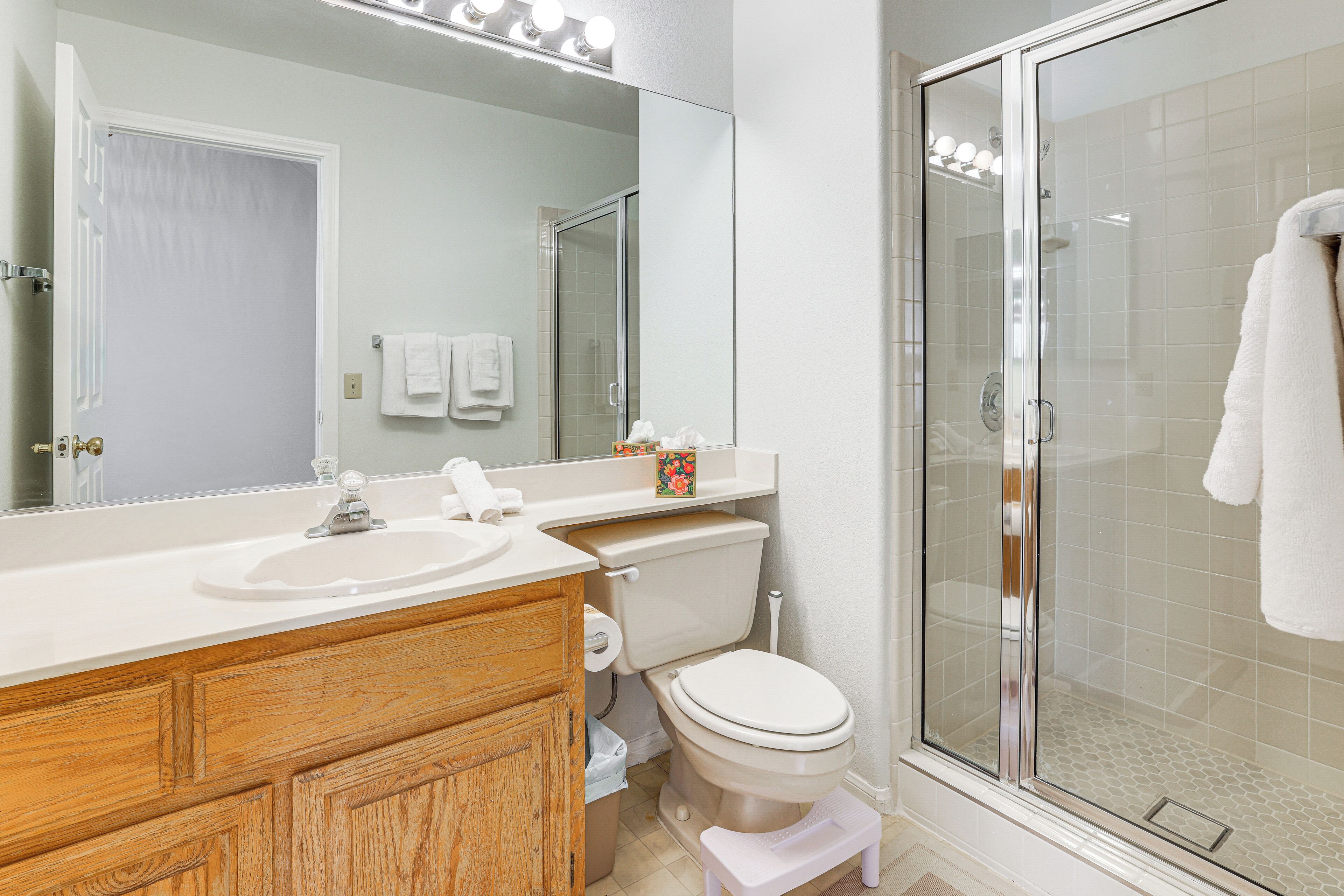 A bathroom with a wooden vanity, a toilet, a glass shower enclosure, and a large mirror with lights above.