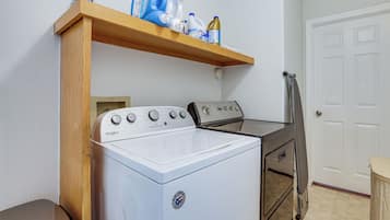 Uma lavanderia com máquina de lavar e secadora Whirlpool, uma prateleira de madeira com produtos de limpeza e uma porta com porta-toalhas.