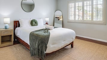 Een net opgemaakt bed met witte lakens, een groene plaid en een houten hoofdbord. Aan de muur boven het bed hangt een ronde spiegel, en bij het bed staat een nachtkastje met een lamp en een klein klokje. De kamer heeft een groot raam met jaloezieën, en de vloer is belegd met een beige vloerkleed.