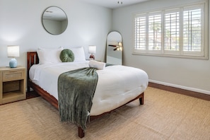 A neatly made bed with white linens, a green throw blanket, and a wooden headboard. There is a round mirror on the wall above the bed, and a bedside table with a lamp and a small clock. The room has a large window with blinds, and the floor is covered with a beige carpet.