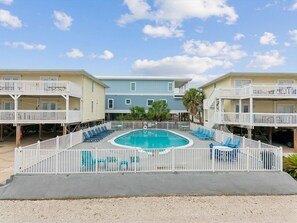 A pool area with a rectangular pool, surrounded by a white fence, and two-story buildings with balconies. - Moonraker #2 | Cozy Condo with Community Pool + Grilling Area (Gulf Shores)