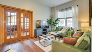 Un salon avec un canapé vert, une table basse, un téléviseur et une grande fenêtre équipée de stores.