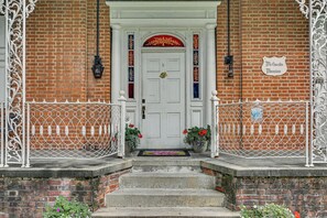 A white door with a red and gold decorative arch, flanked by white iron gates and a brick wall. - 2-apartment Combo in Dtwn Bedford: Hike & Golf! (Bedford)