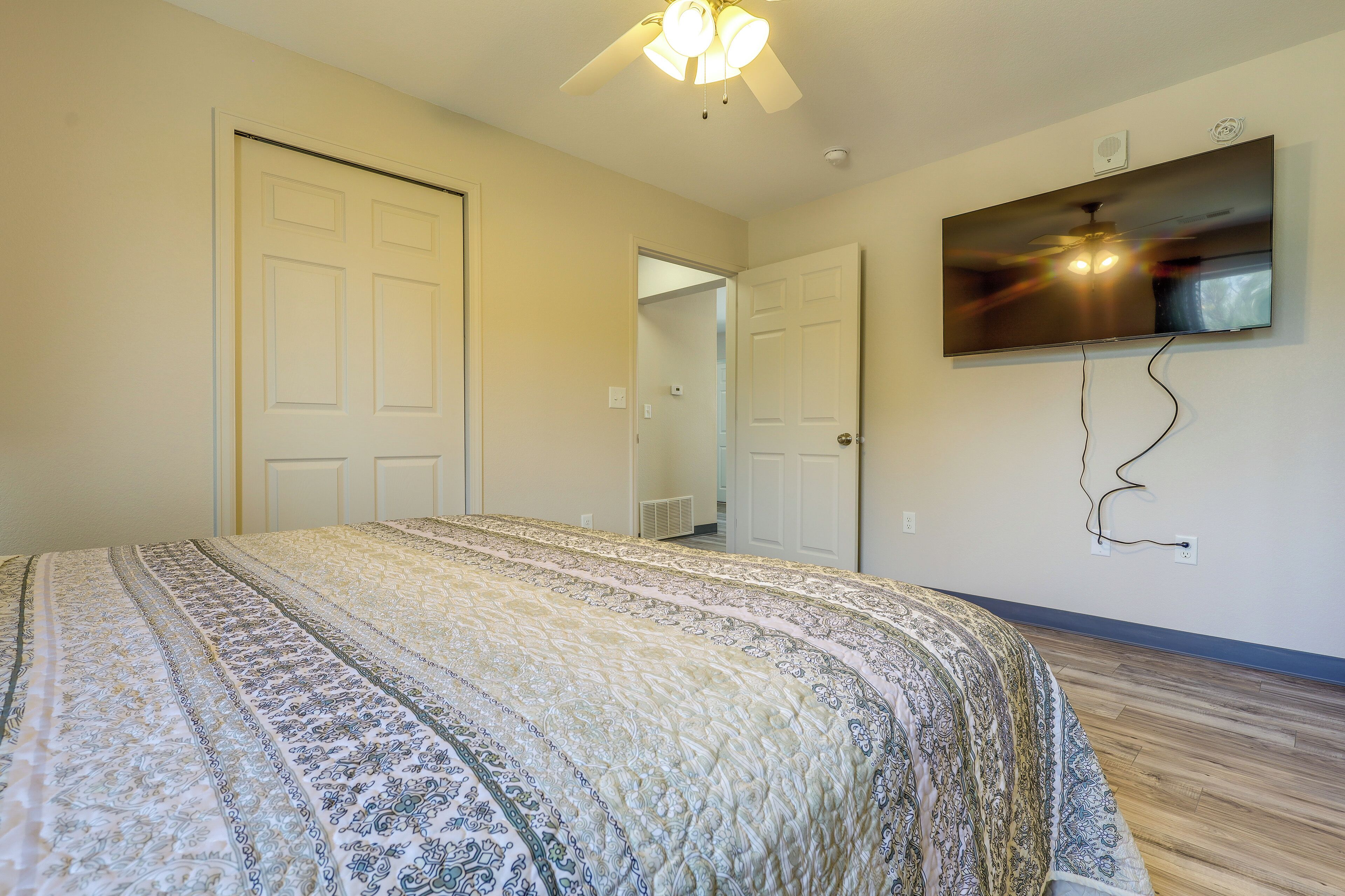A bedroom with a large bed, a ceiling fan, a flat-screen TV mounted on the wall, and a door leading to another room.