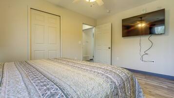 A bedroom with a large bed, a ceiling fan, a flat-screen TV mounted on the wall, and a door leading to another room.