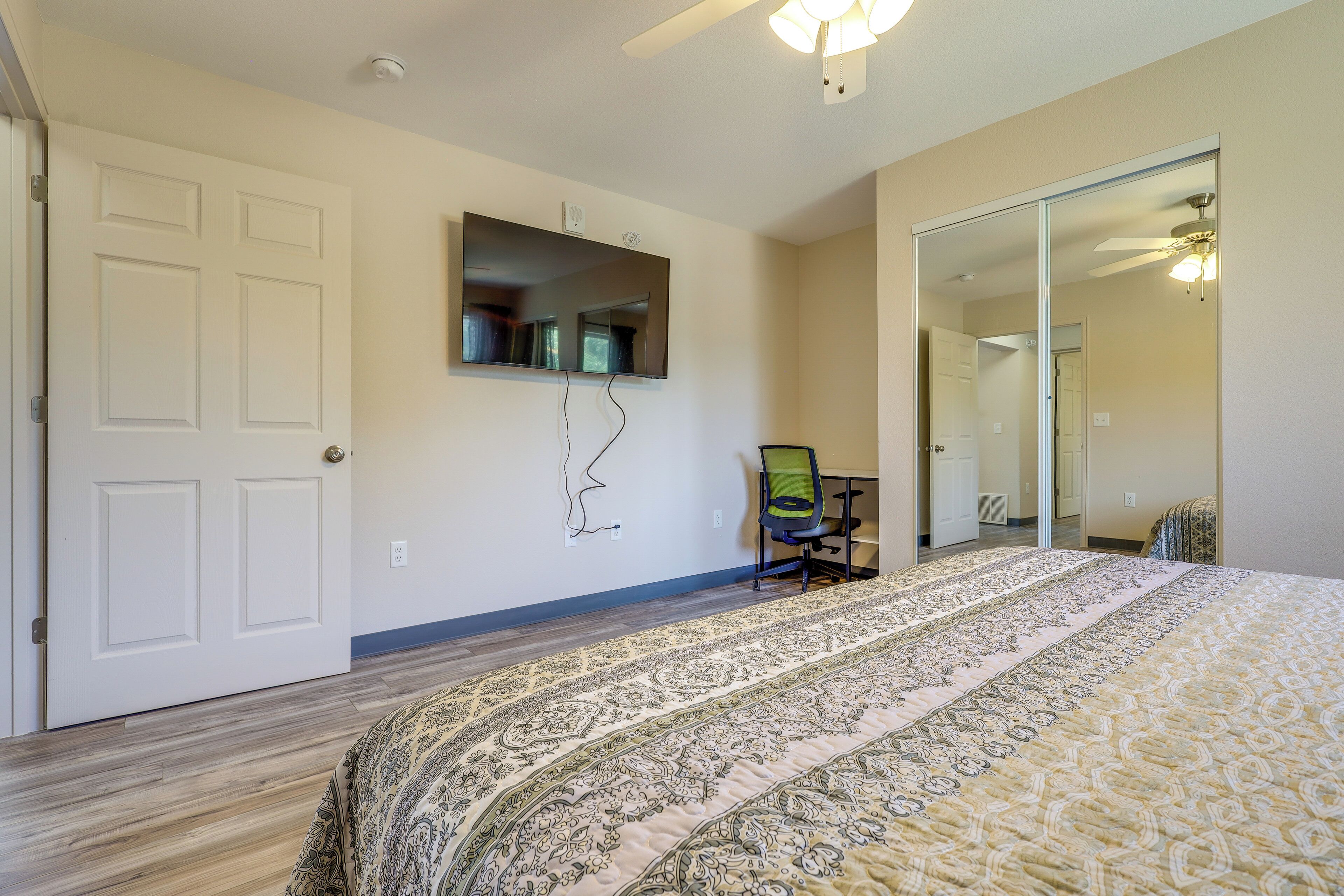A bedroom with a large bed, a flat-screen TV mounted on the wall, a chair, and a door leading to another room.