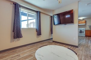 A room with a large window, a flat-screen TV mounted on the wall, and a kitchen area in the background.