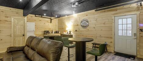 A cozy interior with a leather sofa, a round table, and green chairs. A kitchen area is visible with a refrigerator and various appliances.