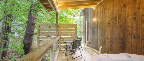 A wooden deck with a hot tub, a table, and chairs, surrounded by trees.