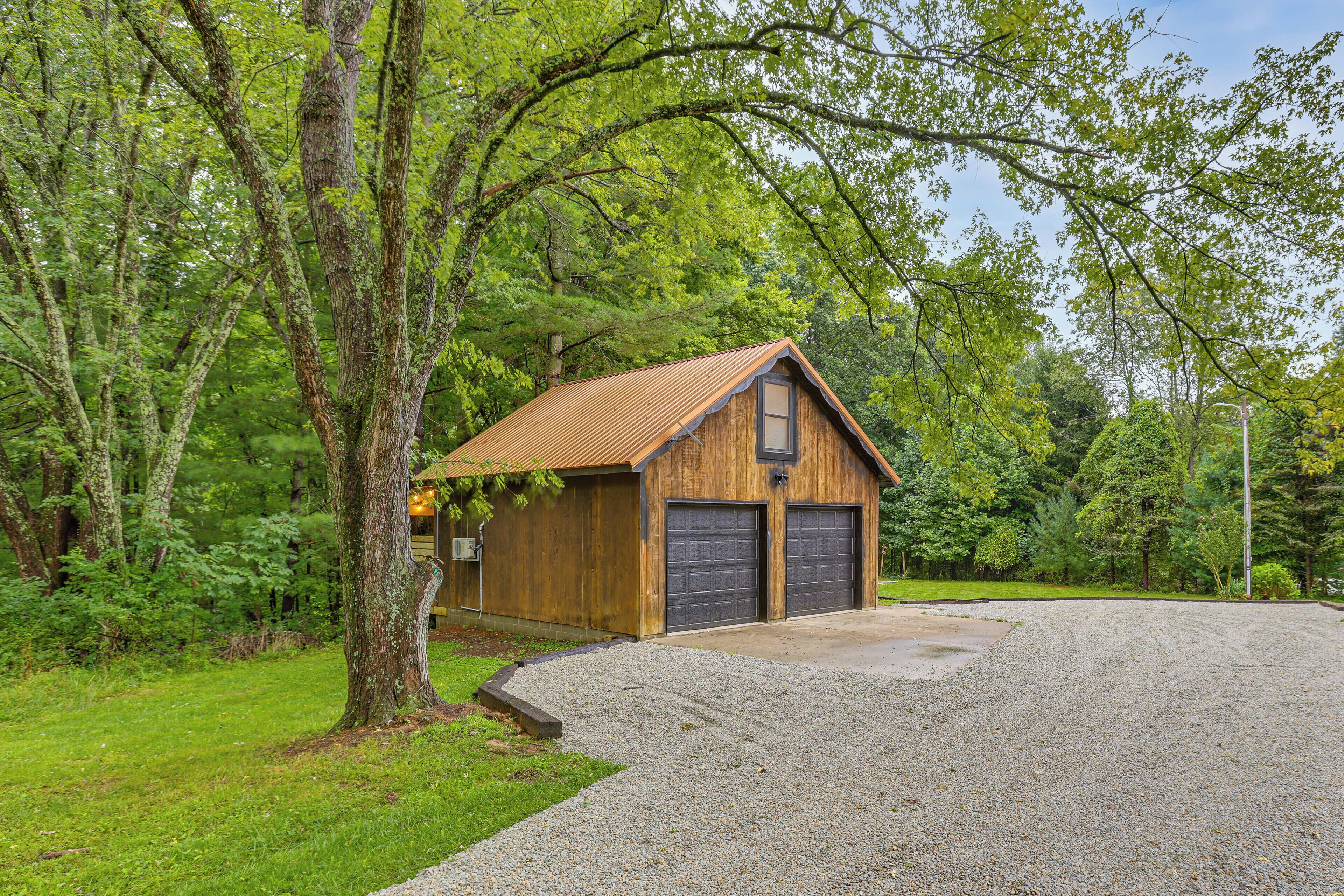 A rustic wooden cabin with a metal roof, surrounded by lush greenery and a gravel driveway.