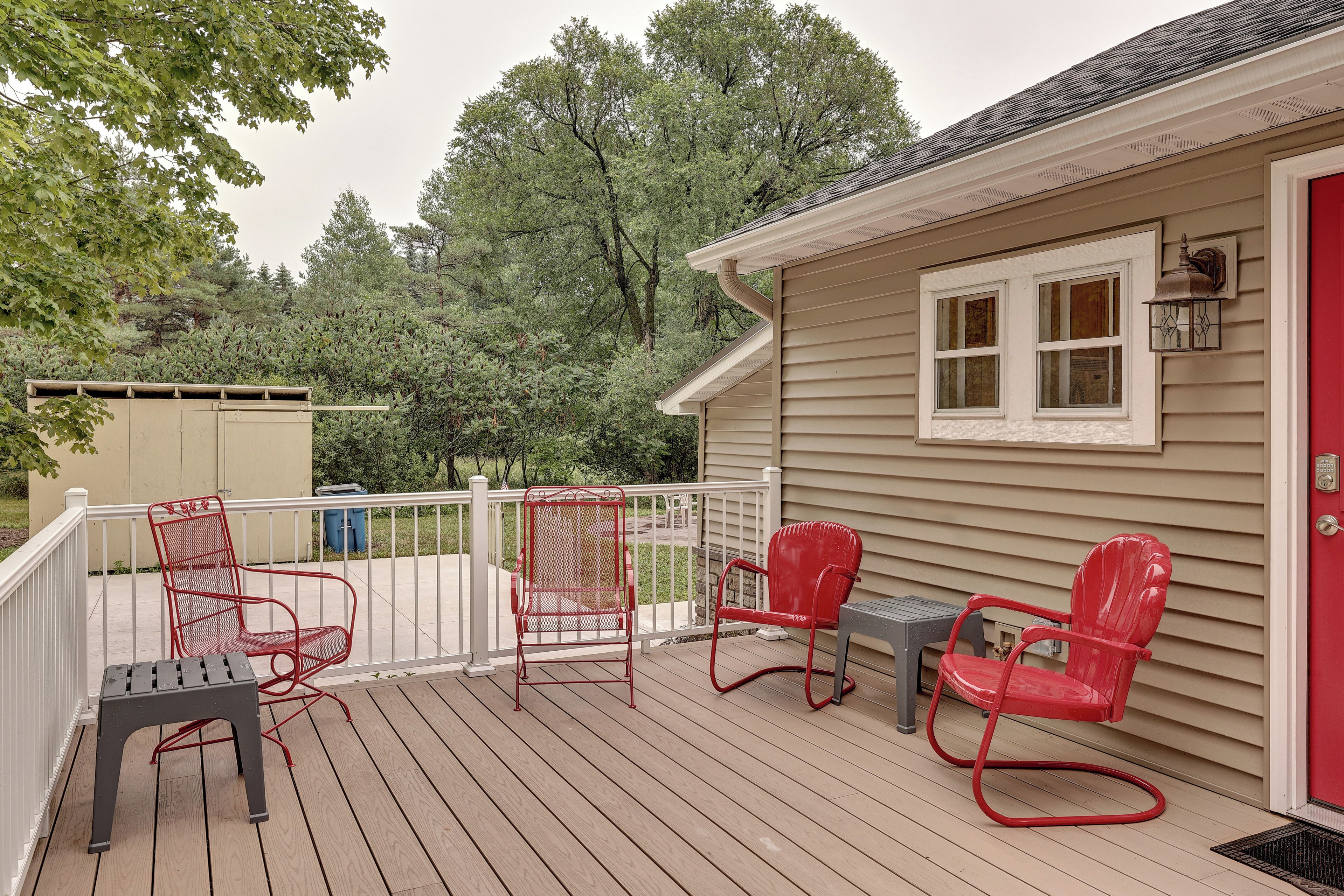 Una terraza techada con sillas metálicas rojas y una mesa, una casa color beige con puerta roja, y una barandilla blanca.