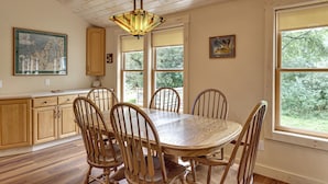 A dining area with a wooden table and chairs, a hanging light fixture, and a large window with a view of greenery. - Golf, Beaches, Slopes & More! Thompsonville Home (Thompsonville)