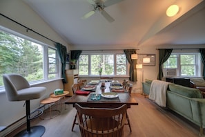 A dining area with a wooden table set for four, surrounded by chairs, with a view of greenery outside.