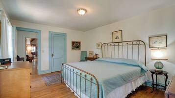 A bedroom with a bed, a dresser, a nightstand, and a lamp. There is a door leading to another room and a window with curtains.