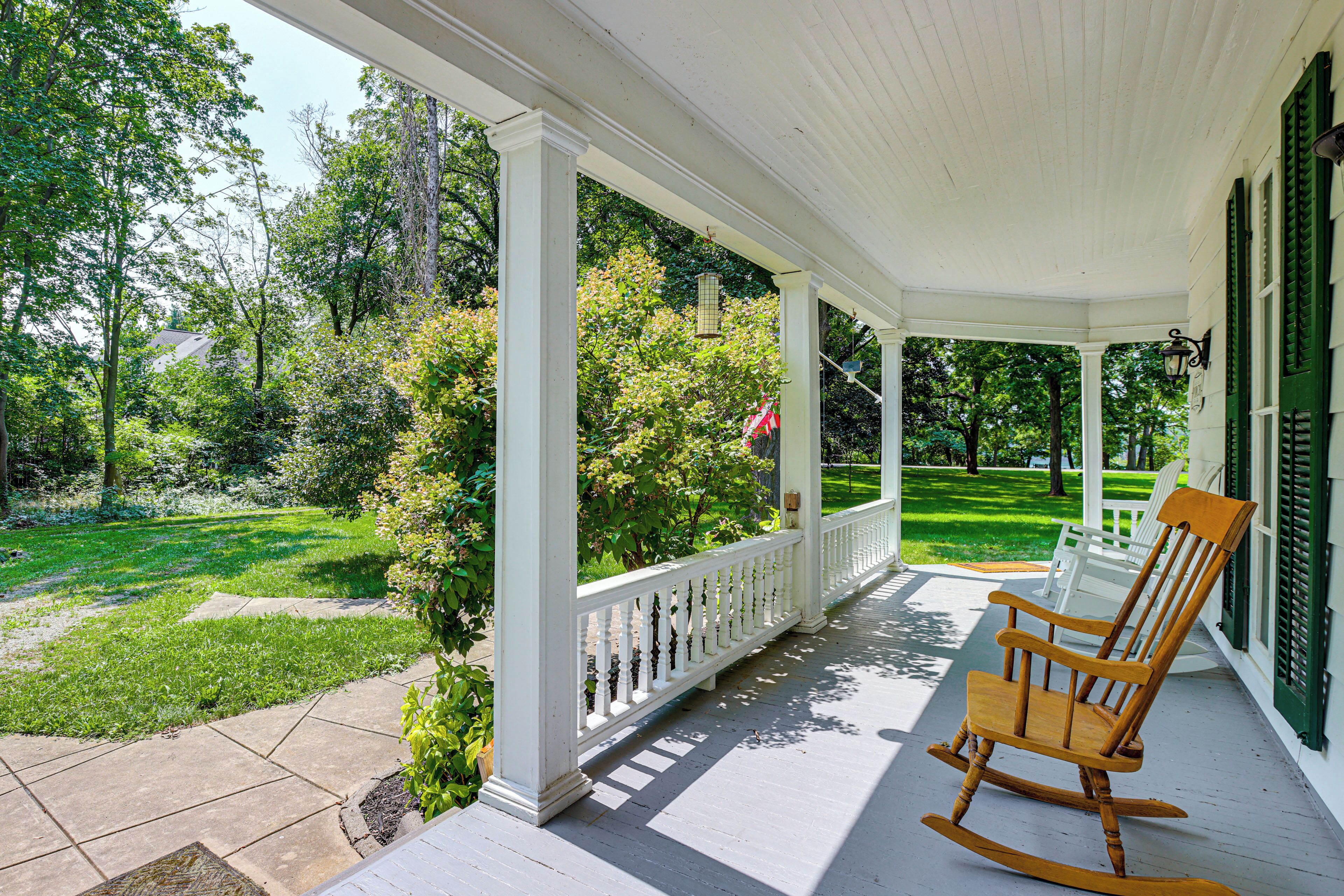 A covered porch with a rocking chair, white railings, and a view of a well-maintained lawn and trees.