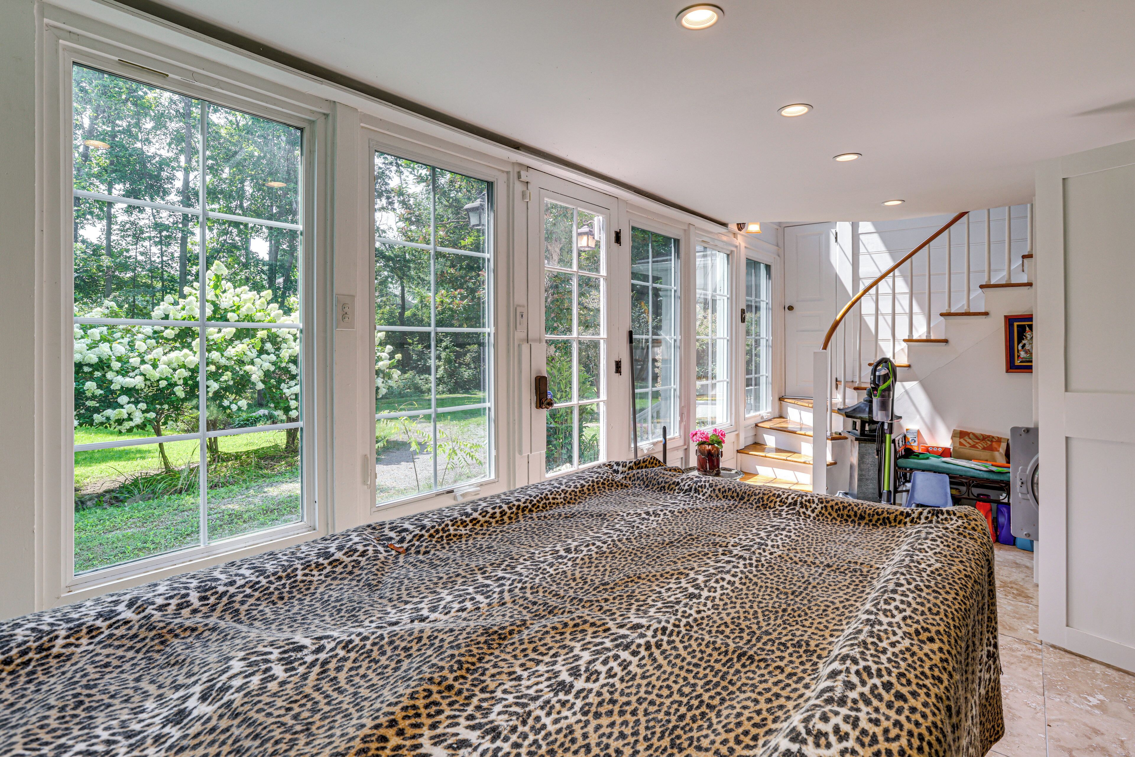 Ett rymligt rum med stora fönster, ett sängöverkast med leopardmönster och utsikt över en trädgård.