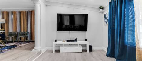 Un salon avec un téléviseur à écran plat fixé au mur, un meuble de rangement blanc et un rideau bleu.