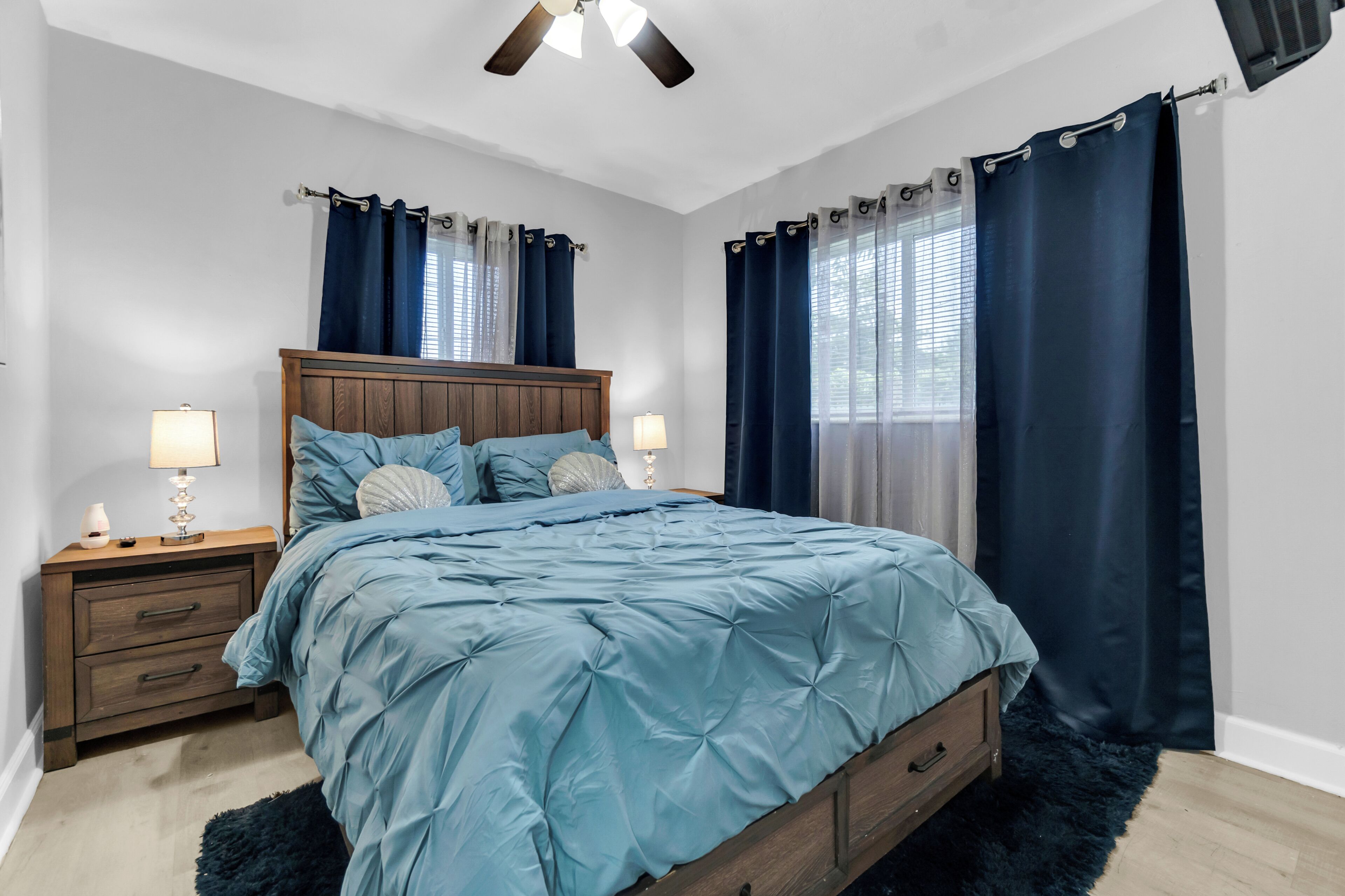 Un dormitorio con cabecera de madera, cama con ropa de cama azul, dos mesitas de noche con lámparas y un ventilador de techo.