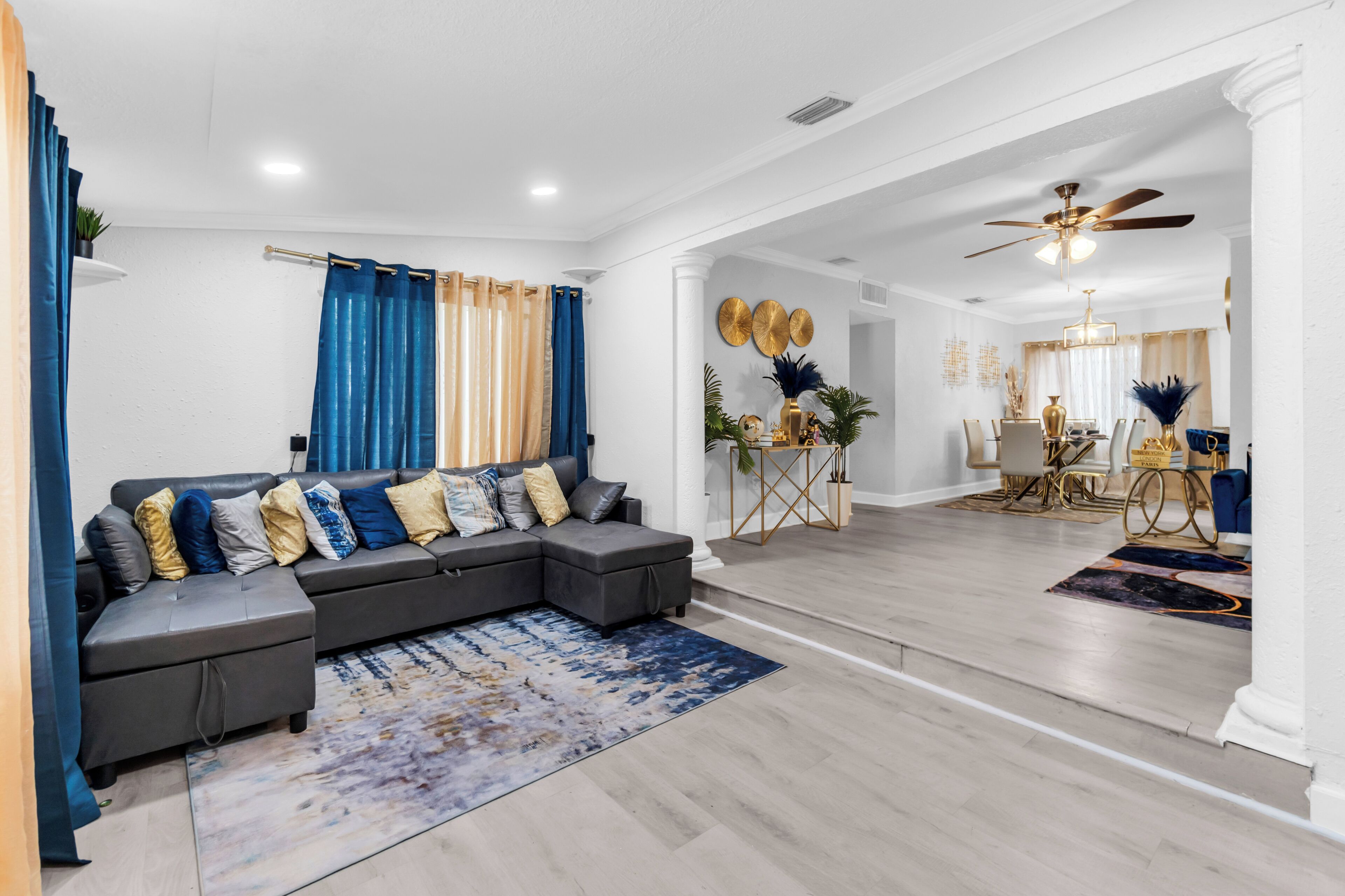 A modern living room with a sectional sofa, a dining area with a chandelier, and a ceiling fan.