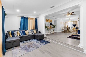 Uma sala de estar moderna com um sofá seccional, uma área de jantar com lustre e um ventilador de teto.