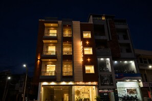Un hotel de varios pisos con ventanas y balcones iluminados, una entrada visible y un letrero que indica 'HOTEL GOD GIFT TV'.