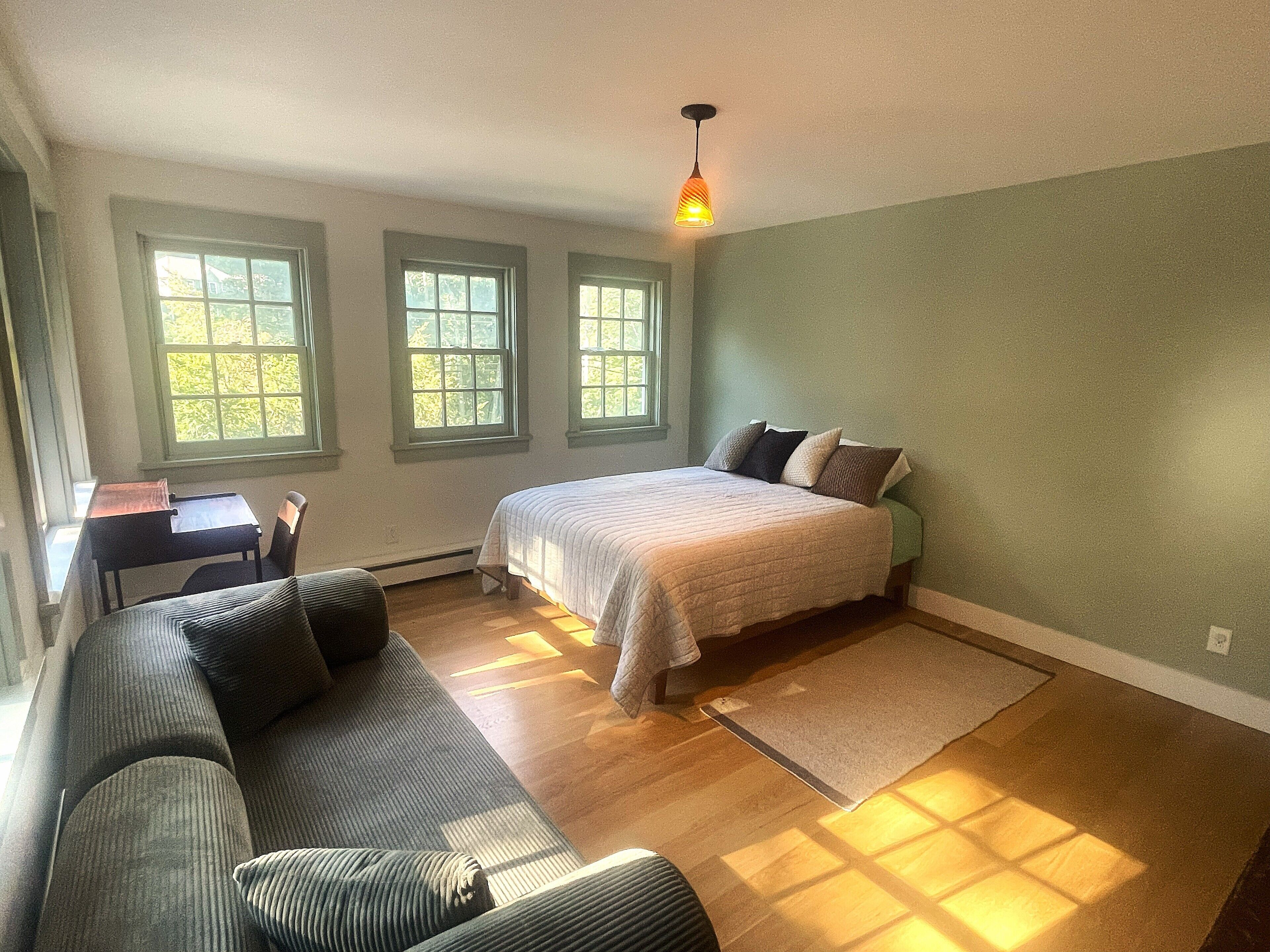 Un dormitorio con cama, sofá y escritorio. Tres ventanas permiten la entrada de luz natural.