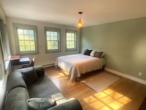 A bedroom with a bed, a sofa, and a desk. There are three windows providing natural light.
