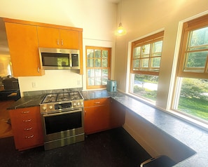 A kitchen with a stove, microwave, and cabinets. There are windows with a view of greenery outside.