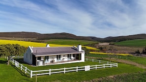 A single-story house with a white exterior and a grey roof, surrounded by a white fence, set against a backdrop of rolling hills and a field of yellow flowers. - Landmeterskop (Stanford)
