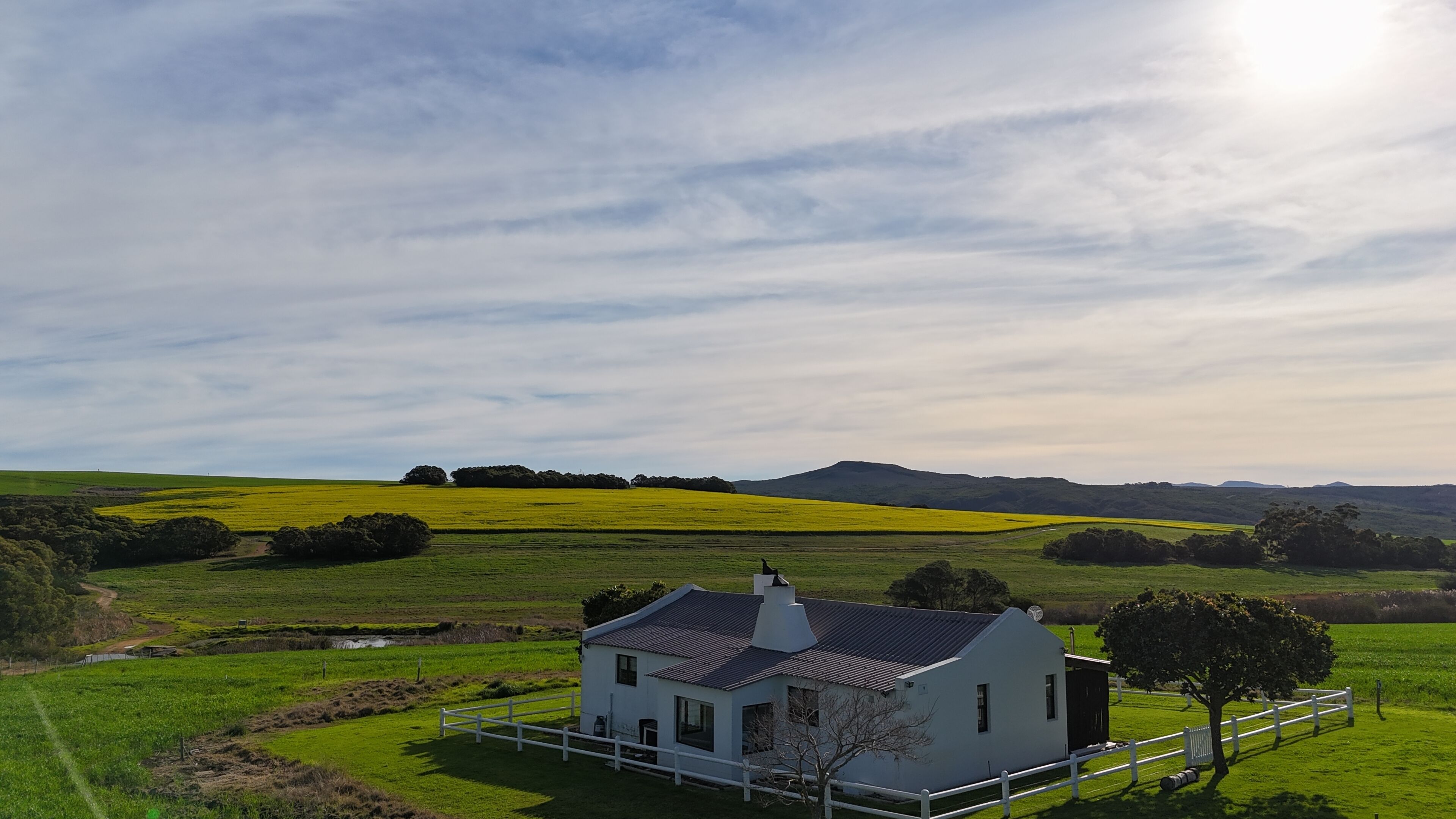 Una casa blanca con techo oscuro, rodeada de campos verdes y un cielo despejado.