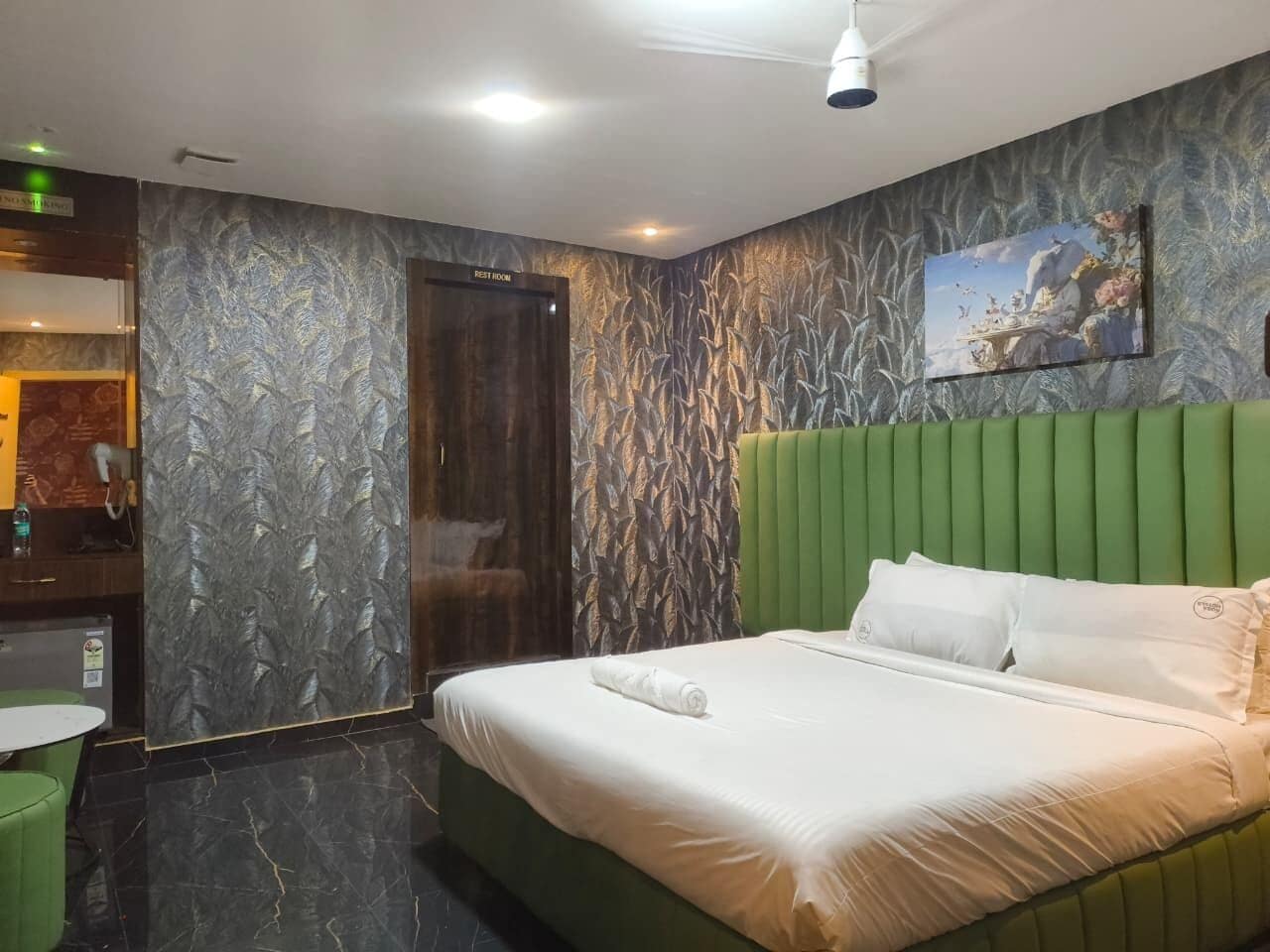 A hotel room with a bed, a green headboard, a painting on the wall, and a bathroom with a mirror and a towel rack.