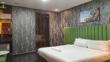 A hotel room with a bed, a green headboard, a painting on the wall, and a bathroom with a mirror and a towel rack.