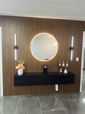 Un baño moderno con pared de paneles de madera, un espejo redondo con iluminación integrada, un mueble de tocador negro y un jarrón decorativo con flores.