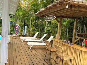 Terraza de madera con sillones de descanso y zona de bar, rodeada de exuberante vegetación tropical.