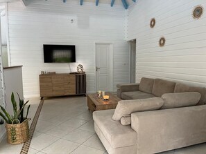 Una sala de estar con un sofá beige, un mueble de madera, un televisor montado en la pared y apliques decorativos.