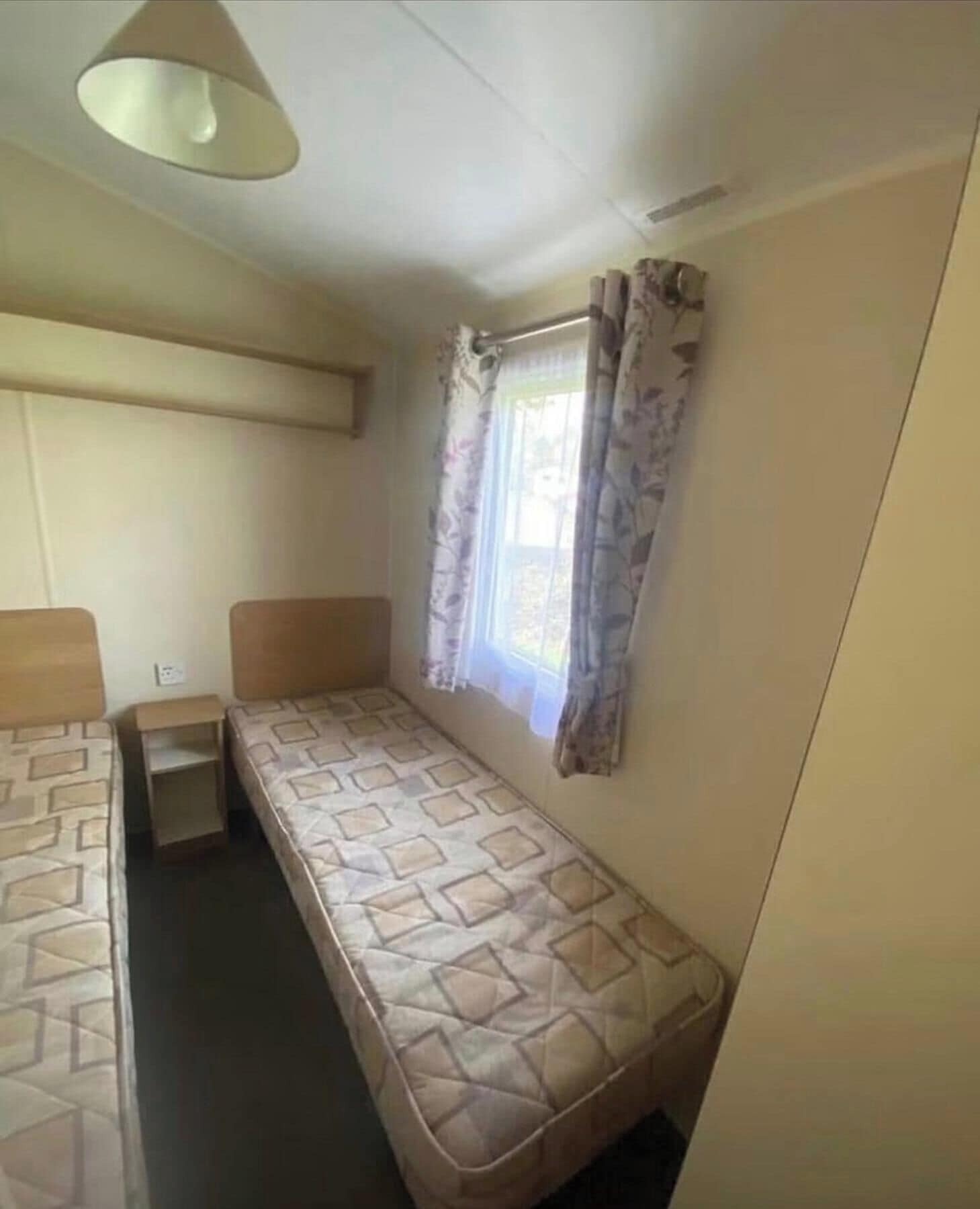 A single-bed room with a window, a bedside table, and a hanging light fixture.