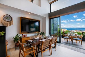 A modern dining area with a large TV, wicker furniture, and a view of the city. - Romantic Zone 2BR Penthouse | Ocean Views & Pool (Puerto Vallarta)
