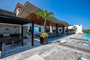 Un espace piscine extérieur moderne, doté de chaises longues, d’un barbecue et d’un patio couvert.