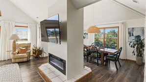 A modern living room with a flat-screen TV mounted on the wall, a fireplace, a dining area with a table and chairs, and a large window offering a view of the outdoors. - New Listing, Private Pool, Hot Tub, Walk to Beach, Dogs Welcome C0919 (Corolla)
