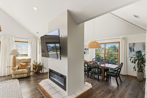 A modern living room with a flat-screen TV mounted on the wall, a fireplace, a dining area with a table and chairs, and a large window offering a view of the outdoors. - New Listing, Private Pool, Hot Tub, Walk to Beach, Dogs Welcome C0919 (Corolla)