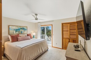 Ein Schlafzimmer mit einem Bett, einem Deckenventilator, einem Gemälde, einem Nachttisch, einer Lampe und Blick ins Freie durch eine Schiebetür.