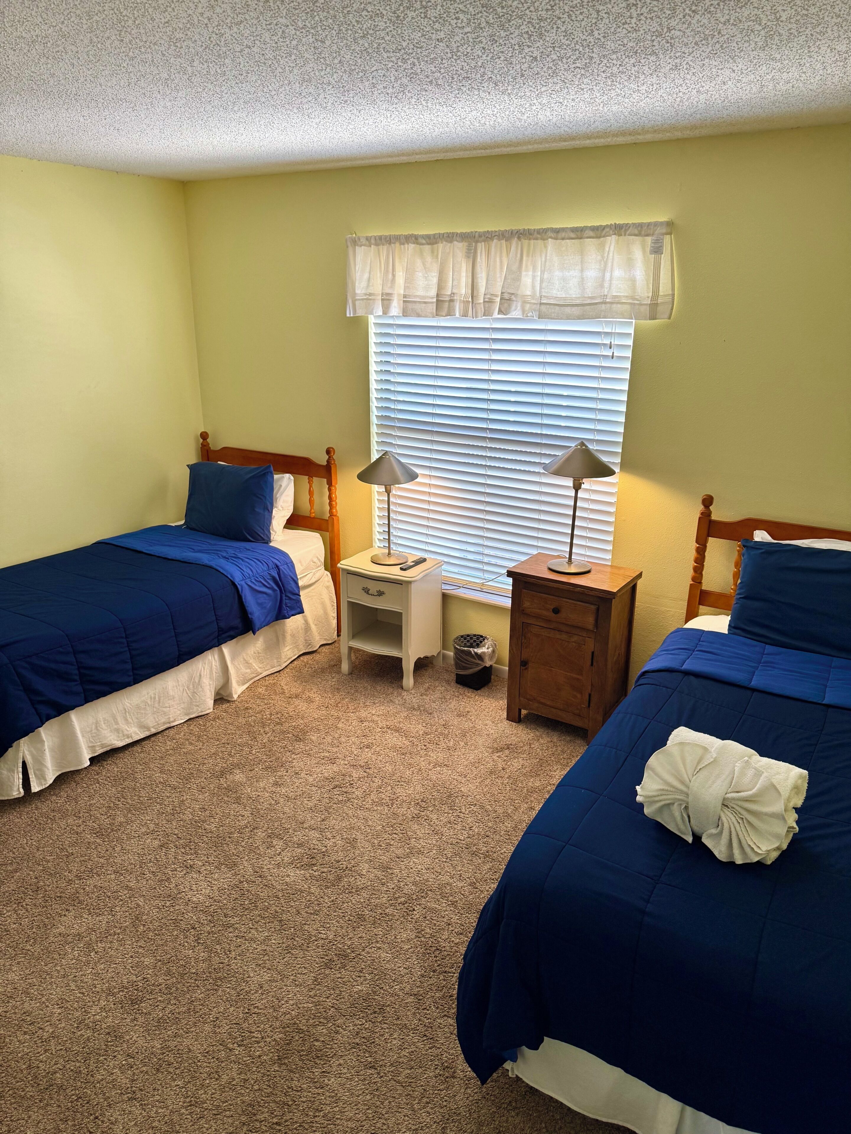 A bedroom with two beds, a nightstand, a lamp, and a window with blinds.