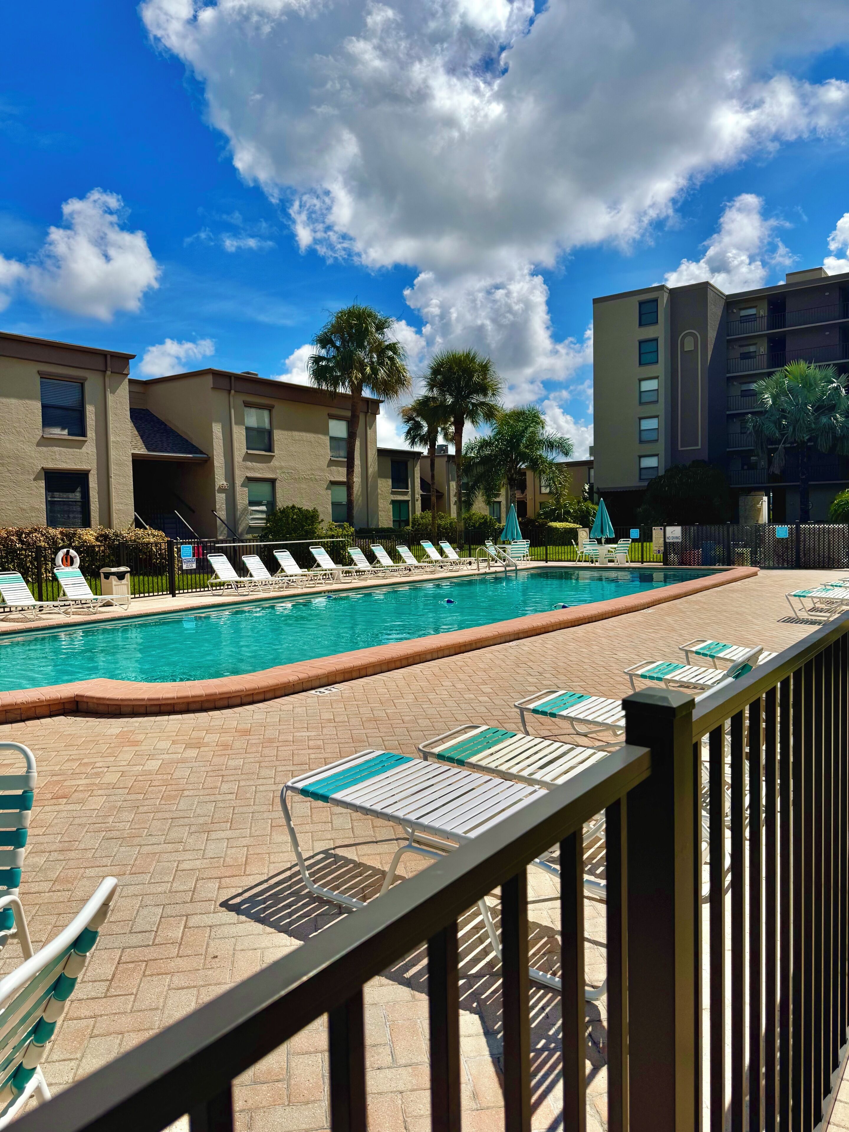 A swimming pool with lounge chairs and a brick deck, surrounded by buildings and palm trees.