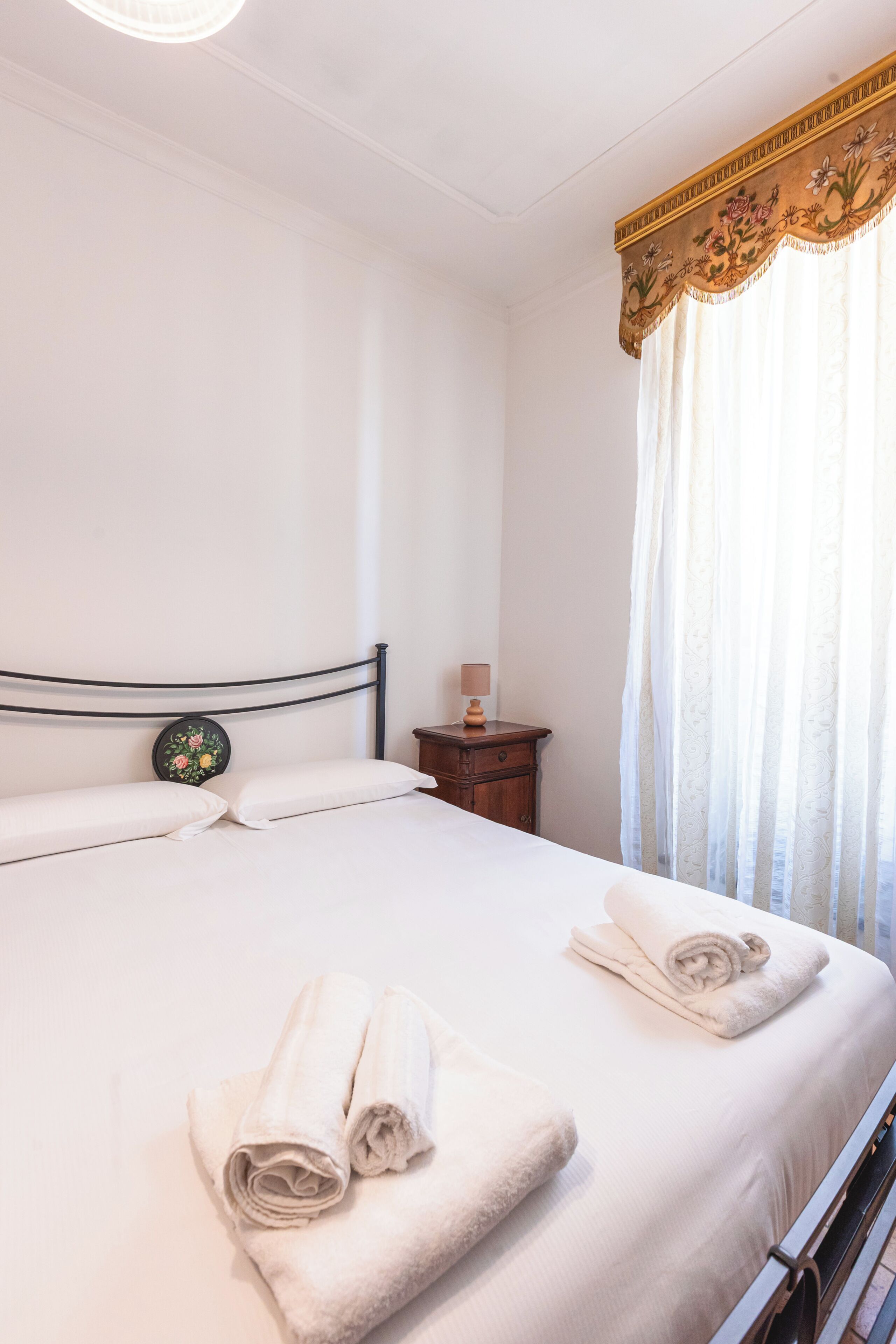 Una camera d'albergo con un letto, cuscini, un comodino, una lampada e una finestra con tende.