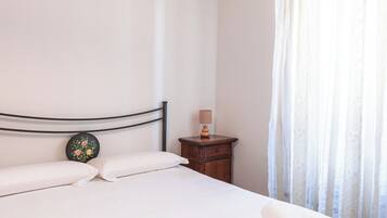 Una camera d'albergo con un letto, cuscini, un comodino, una lampada e una finestra con tende.