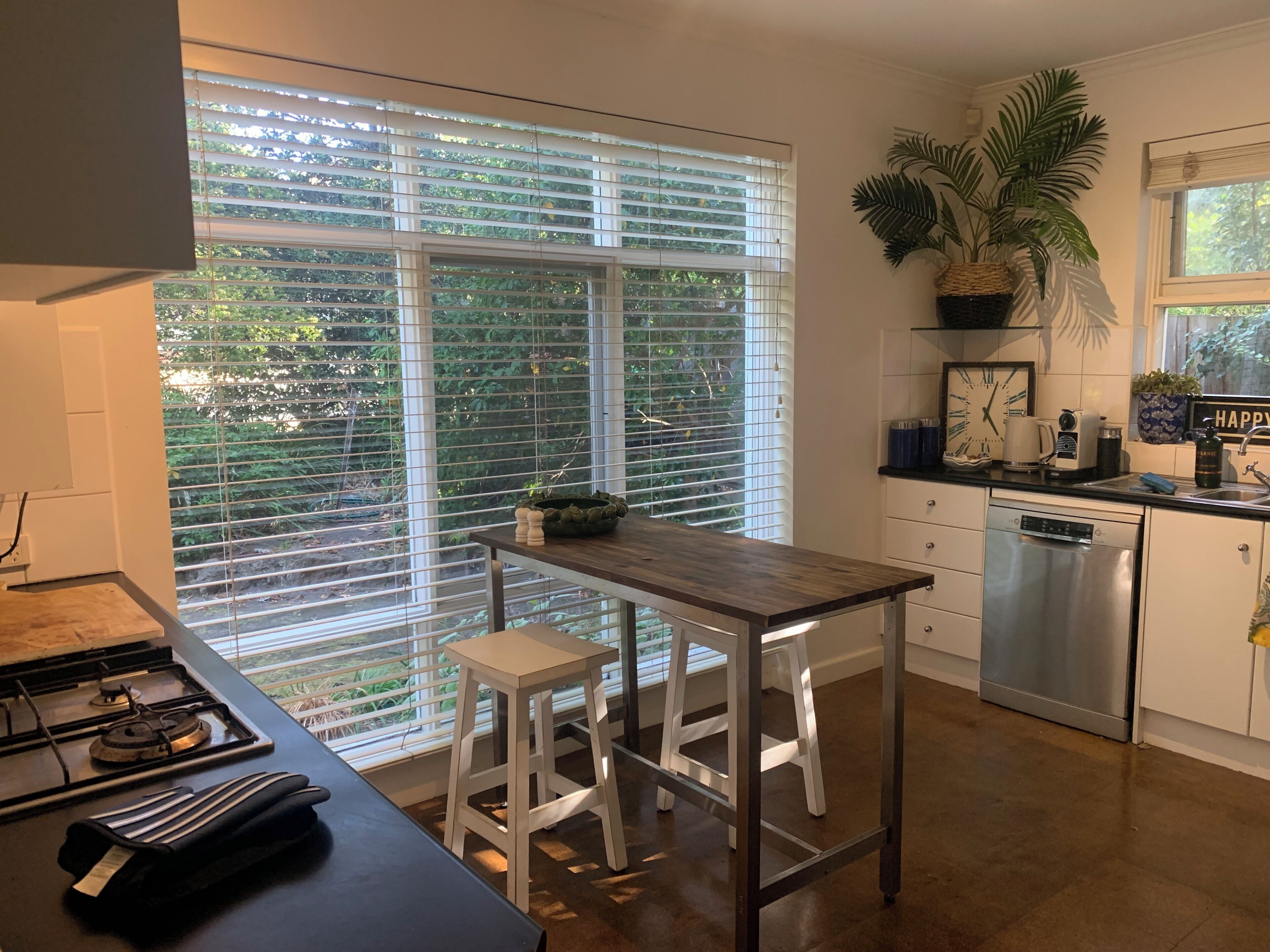 A modern kitchen with a large window, white cabinetry, a stainless steel dishwasher, and a wooden table.