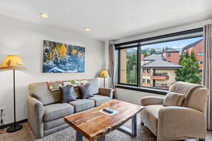 A modern living room with a sofa, armchair, and coffee table. There is a painting on the wall and a window with a view of buildings. - Ski-Inspired Mountain Getaway Steps from Peak 9 and Main Street! (Breckenridge)