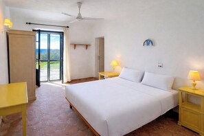 A hotel room with a bed, two bedside tables with lamps, a ceiling fan, a door leading outside, and a small shelf on the wall.