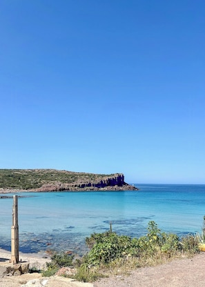 A coastal landscape with clear blue waters, a rocky outcrop, and sparse vegetation.